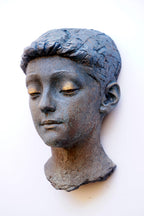A textured sculpture of a young boy's head with closed eyes, mounted on a plain white wall.