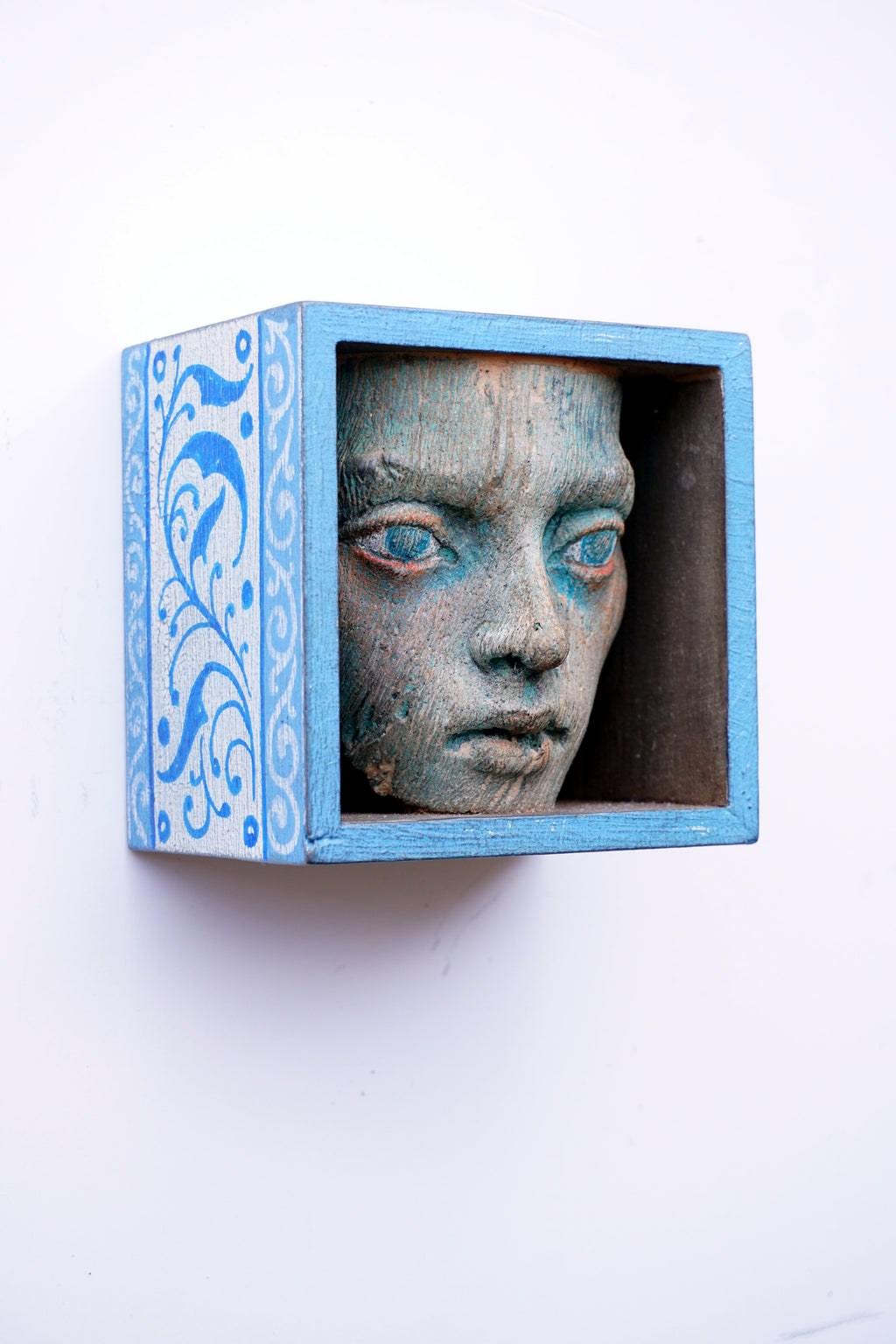 A decorative light blue wooden shadow box mounted on a white wall, containing a textured, weathered stone-like classical female face with blue-painted eyes.