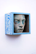 A decorative light blue wooden shadow box mounted on a white wall, containing a textured, weathered stone-like classical female face with blue-painted eyes.