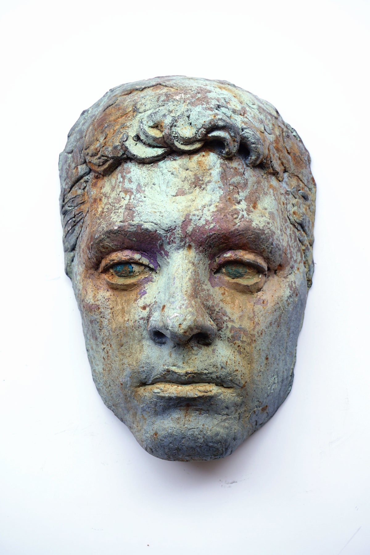 An ancient-style bronze- or metal-like sculpture of a male face with textured patina, photographed against a plain white background.