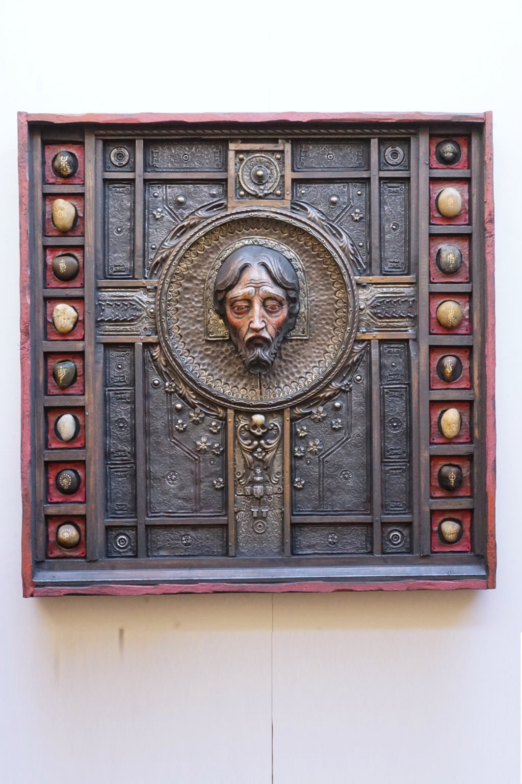 An ornate square religious wall plaque featuring a sculpted bust of Jesus Christ in a circular frame, surrounded by a dark textured panel with decorative medallions and a reddish-brown border, mounted on a plain off-white wall.