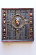 An ornate square religious wall plaque featuring a sculpted bust of Jesus Christ in a circular frame, surrounded by a dark textured panel with decorative medallions and a reddish-brown border, mounted on a plain off-white wall.