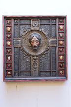 An ornate square religious wall plaque featuring a sculpted bust of Jesus Christ in a circular frame, surrounded by a dark textured panel with decorative medallions and a reddish-brown border, mounted on a plain off-white wall.