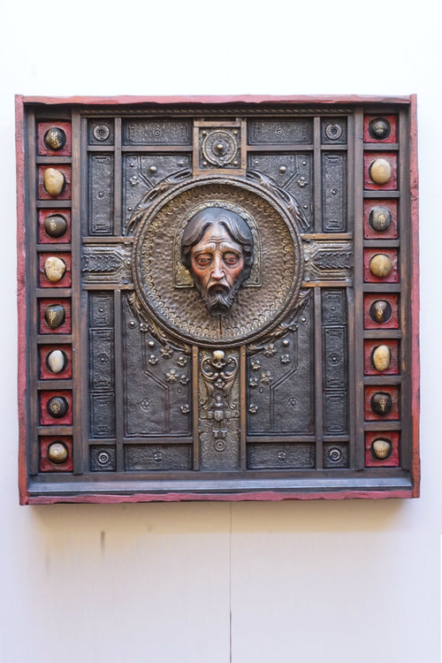 An ornate square religious wall plaque featuring a sculpted bust of Jesus Christ in a circular frame, surrounded by a dark textured panel with decorative medallions and a reddish-brown border, mounted on a plain off-white wall.