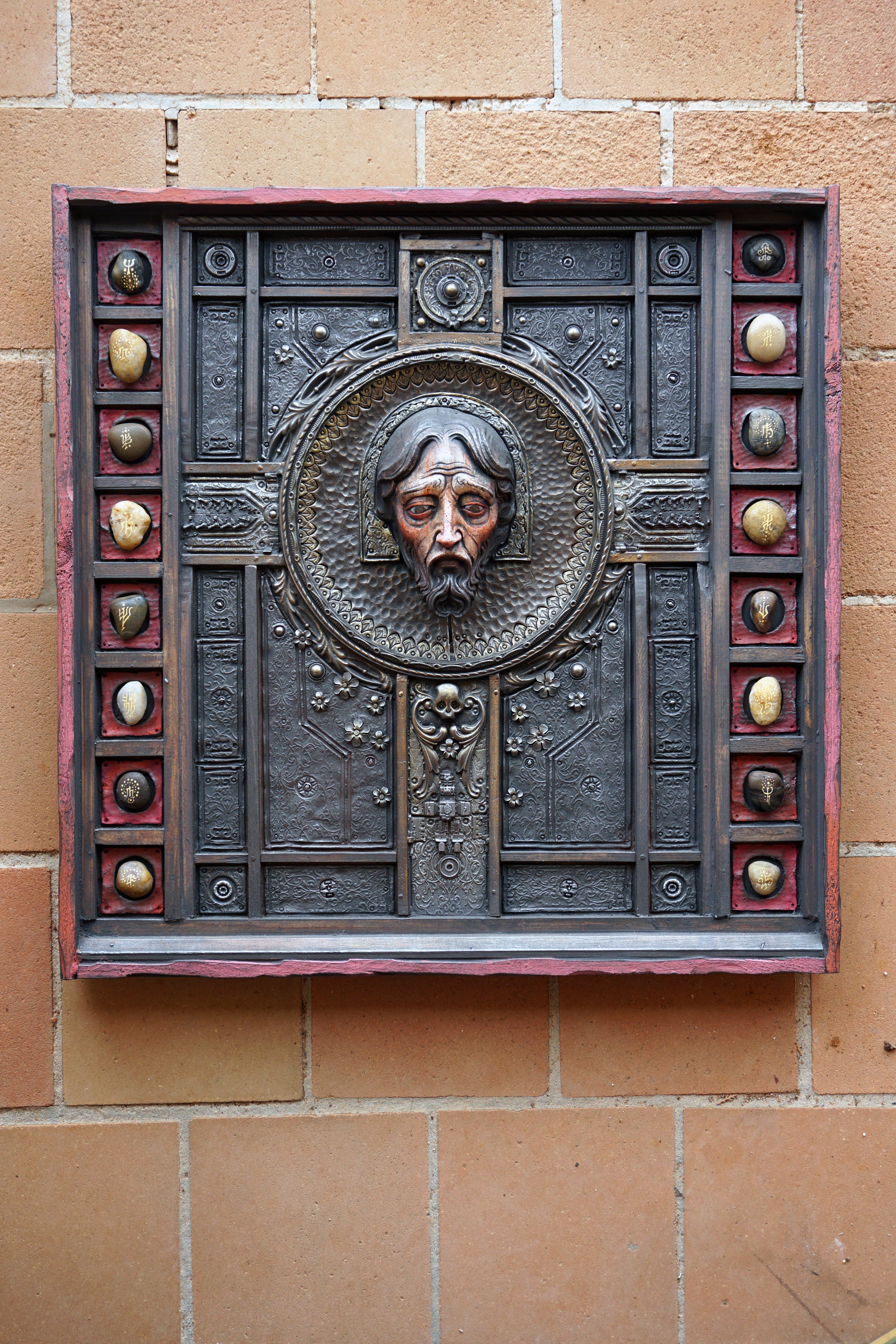 A square dark metal wall plaque featuring a raised relief portrait of Jesus Christ in the center, surrounded by decorative geometric panels and small circular metallic accents, mounted on a tan brick wall.