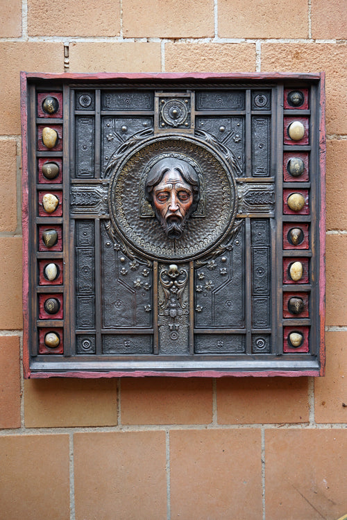 A square dark metal wall plaque featuring a raised relief portrait of Jesus Christ in the center, surrounded by decorative geometric panels and small circular metallic accents, mounted on a tan brick wall.