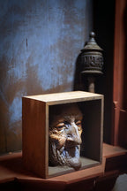 An antique carved wooden box display featuring a sculpted face of an elderly man, set against a dark textured wall with a decorative metal object in the background.