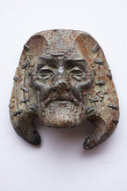 An antique bronze or iron mask with a weathered patina, featuring a stern face with a beard and curved horns, photographed against a plain white background.