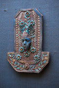 An ornate decorative wall plaque featuring a carved face surrounded by floral and leaf motifs in turquoise and gold tones, mounted on a dark blue-gray textured wall.
