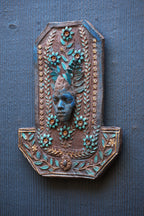 An ornate decorative wall plaque featuring a carved face surrounded by floral and leaf motifs in turquoise and gold tones, mounted on a dark blue-gray textured wall.