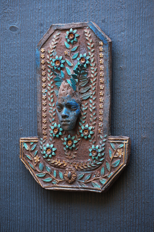 An ornate decorative wall plaque featuring a carved face surrounded by floral and leaf motifs in turquoise and gold tones, mounted on a dark blue-gray textured wall.