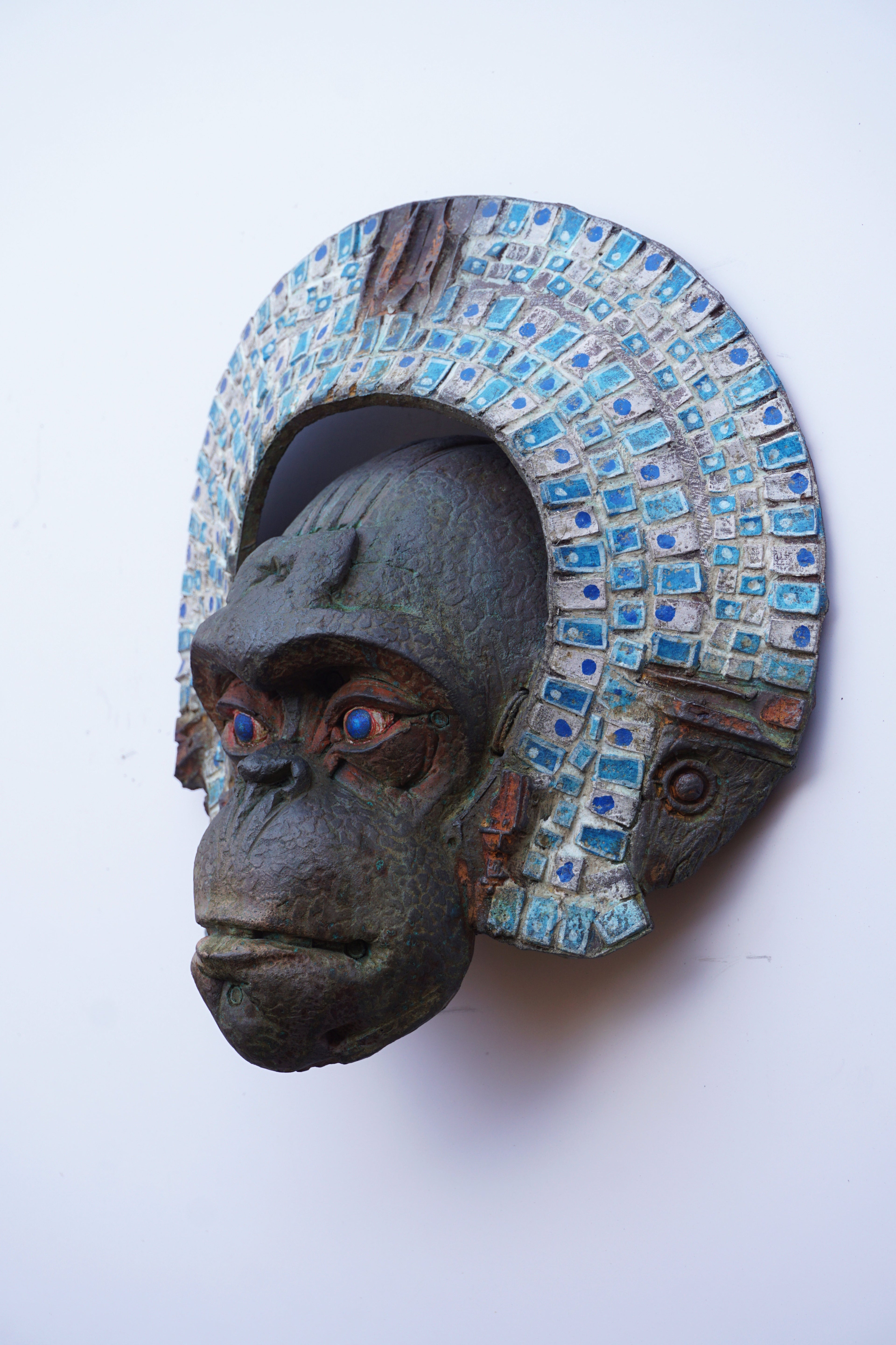 A carved gorilla mask with a textured dark gray face and a large arched headdress made of small blue and white mosaic tiles, shown against a plain white background.