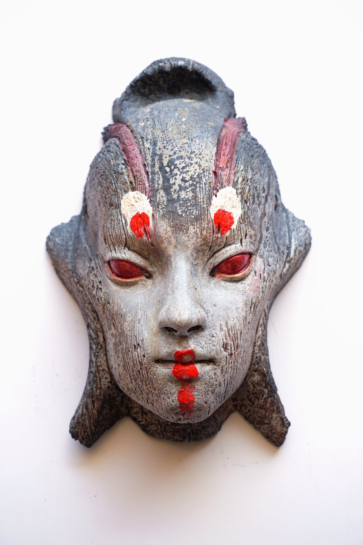 A textured, hand-carved Japanese-style mask with a weathered gray stone-like finish, featuring red eyes, red painted lips, and white and red markings on the forehead, shown against a plain white background.