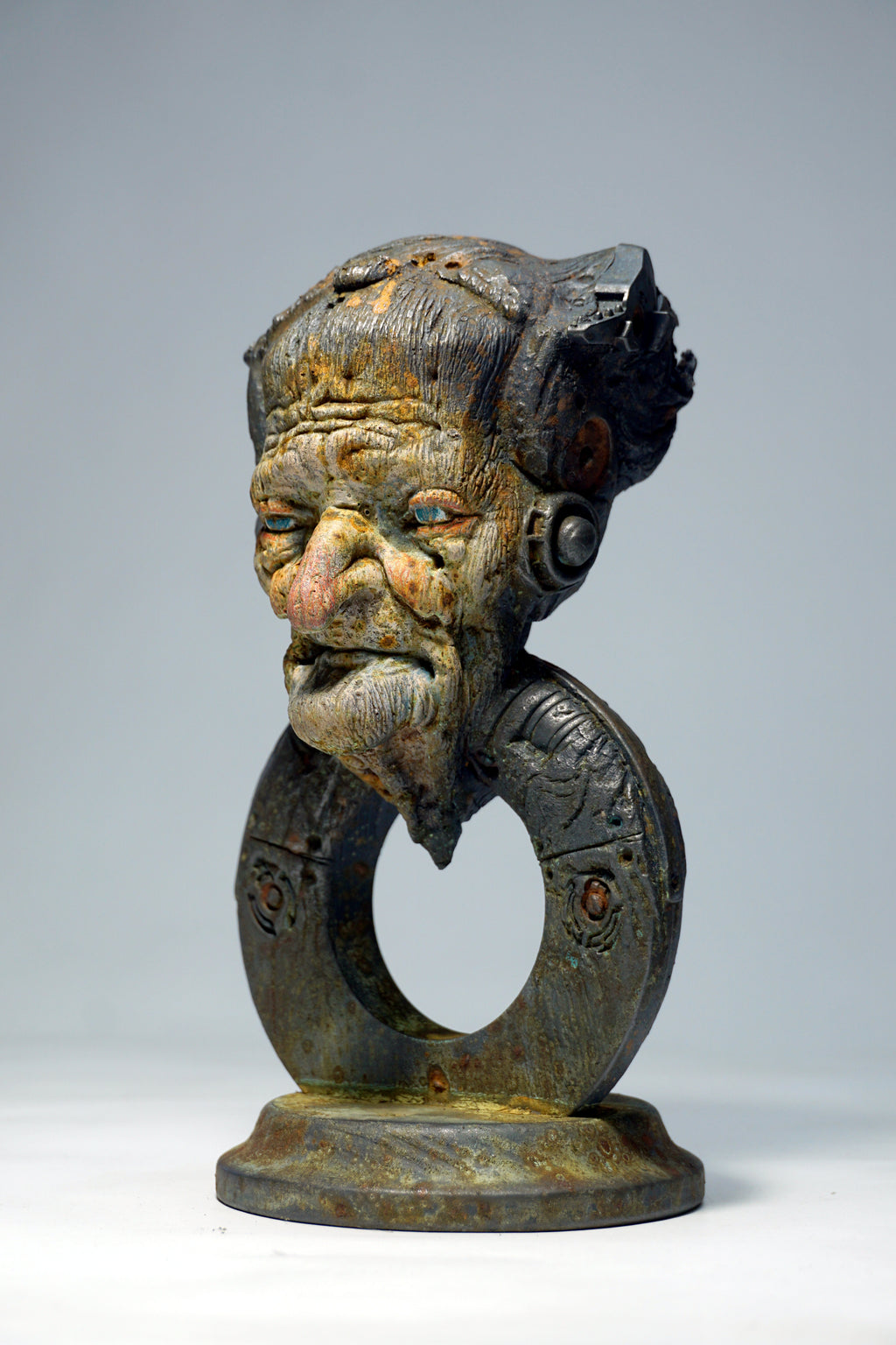 A carved wooden sculpture of a stylized face with a ring-like neck and circular base, shown against a plain light gray studio background.
