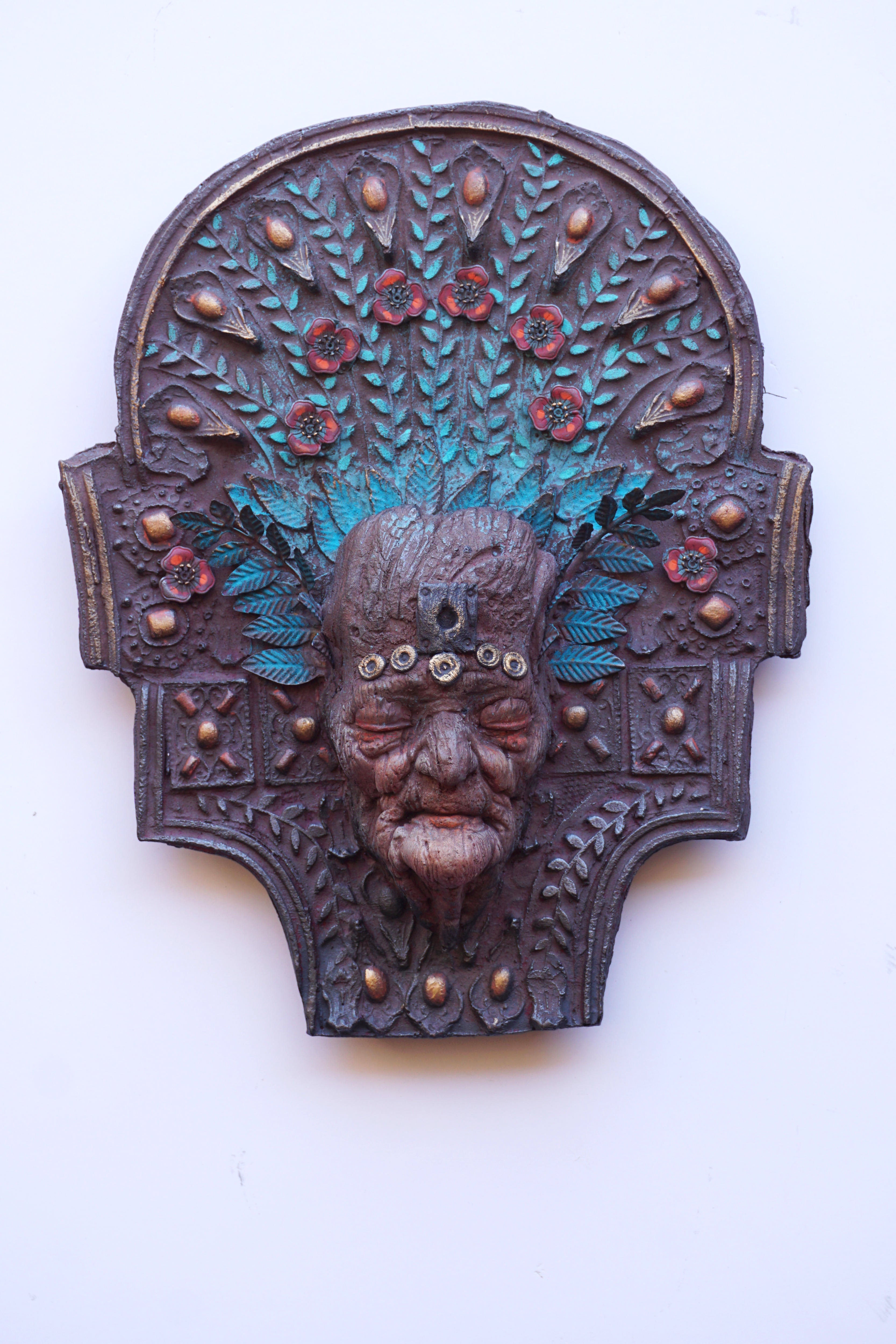An ornate carved wooden-like wall plaque depicting a stylized face with turquoise and red painted details, mounted against a plain white background.