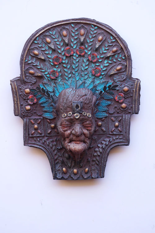 An ornate carved wooden-like wall plaque depicting a stylized face with turquoise and red painted details, mounted against a plain white background.