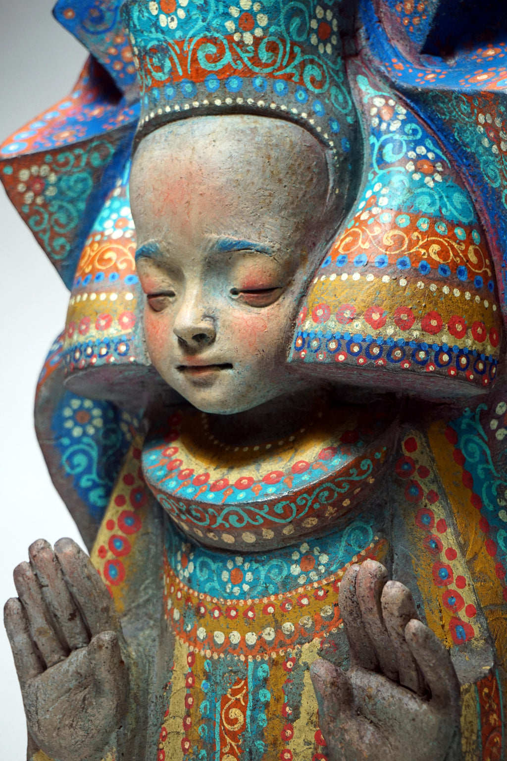 A close-up of a hand-painted wooden figurine of a child with closed eyes, wearing a large decorative headdress and robe decorated with intricate blue, orange, red, and yellow patterns.