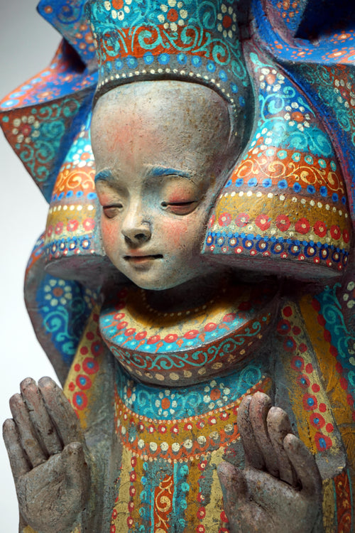 A close-up of a hand-painted wooden figurine of a child with closed eyes, wearing a large decorative headdress and robe decorated with intricate blue, orange, red, and yellow patterns.