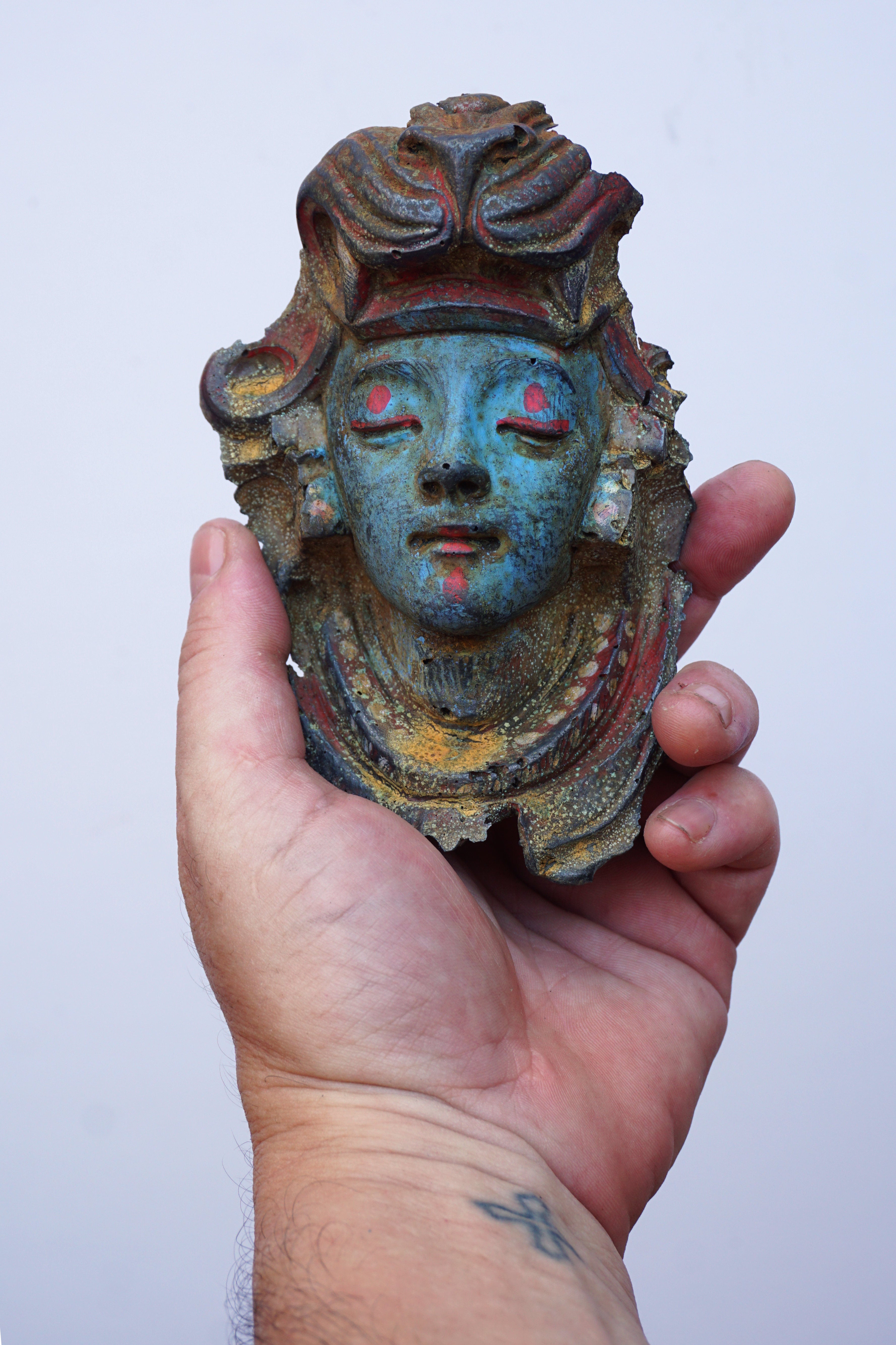 A hand holding a small decorative mask with a blue-green face, red painted details, and aged brown and gold patina against a plain light background.