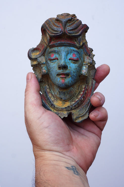 A hand holding a small decorative mask with a blue-green face, red painted details, and aged brown and gold patina against a plain light background.