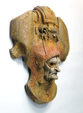 An antique-style bronze and rust-colored metal wall sculpture depicting a stylized helmeted head with a weathered, aged finish, mounted against a plain white wall.
