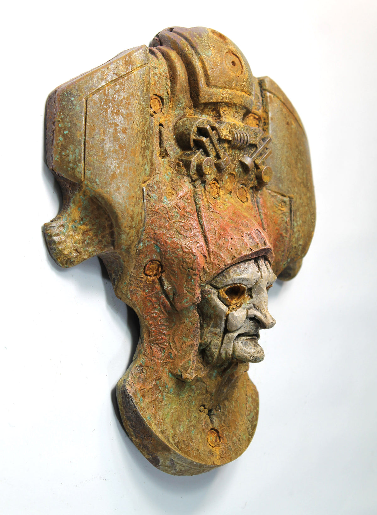 An antique-style bronze and rust-colored metal wall sculpture depicting a stylized helmeted head with a weathered, aged finish, mounted against a plain white wall.