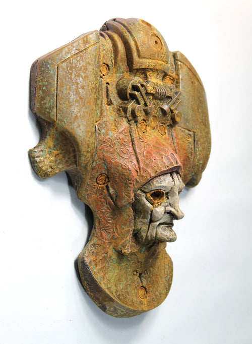 An antique-style bronze and rust-colored metal wall sculpture depicting a stylized helmeted head with a weathered, aged finish, mounted against a plain white wall.