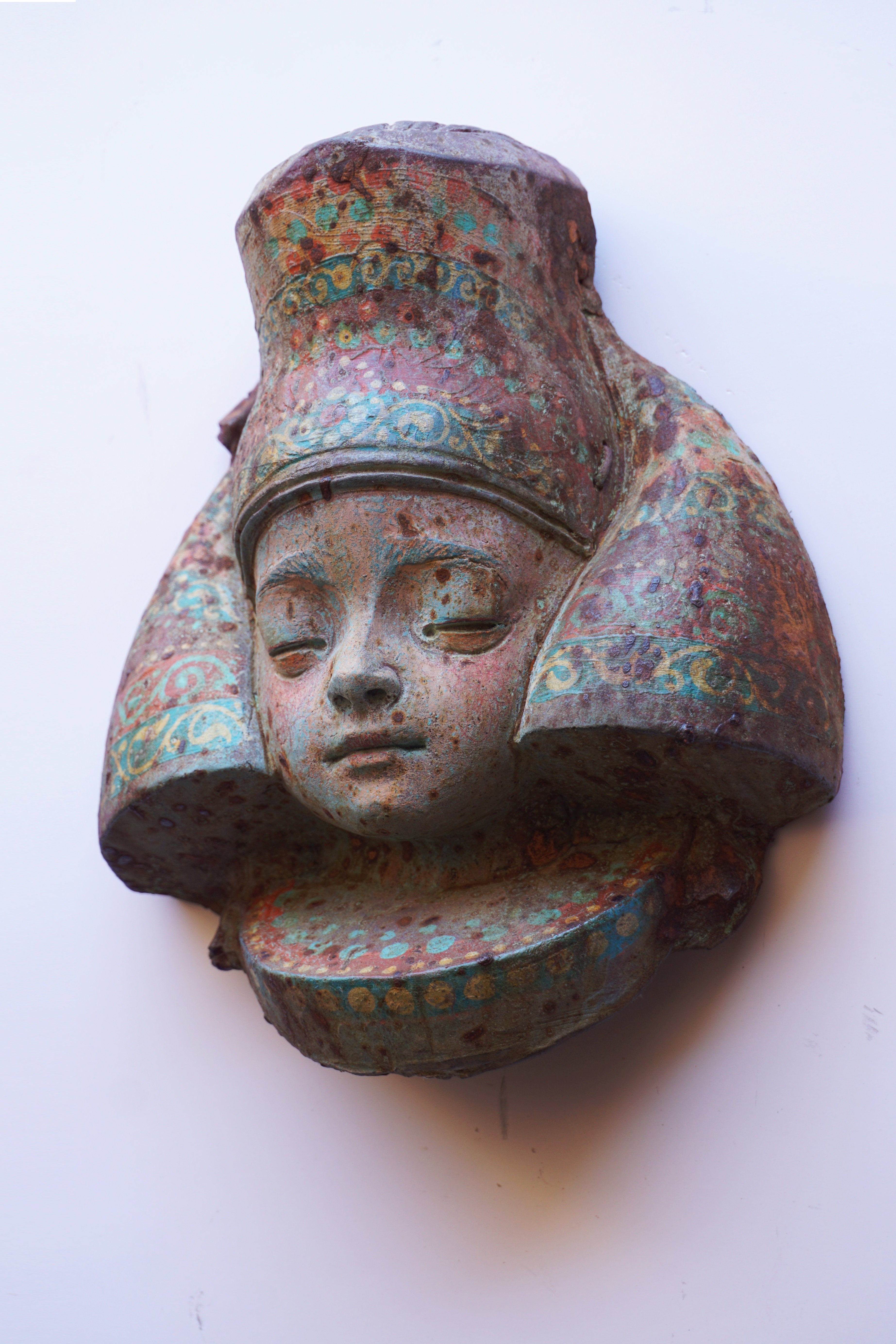 An ancient-style resin wall sculpture depicting a face wearing a tall headdress, with a patina of green, blue, and brown rust-like oxidation, mounted on a plain white wall.