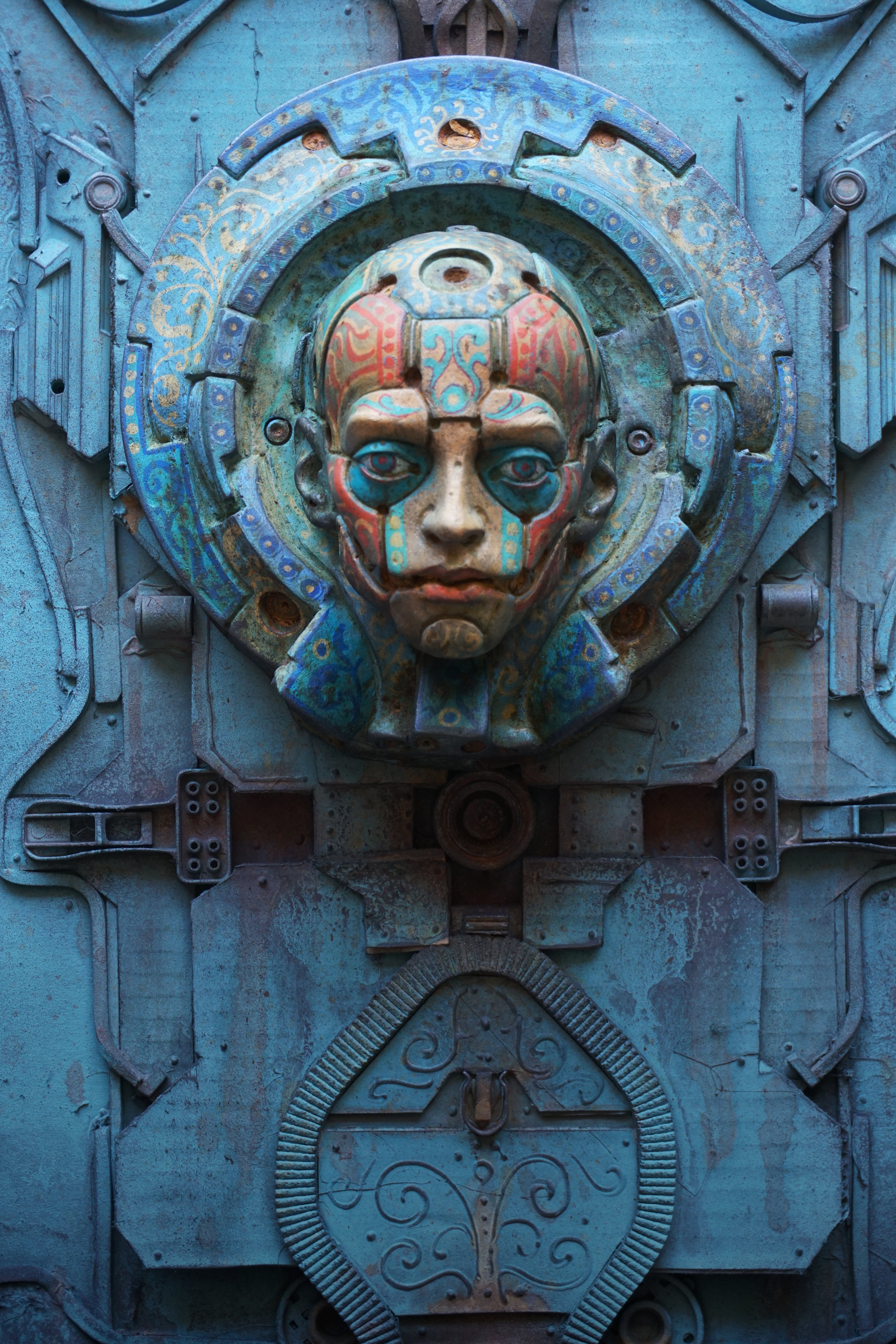 Close-up of an ornate blue metal door featuring a sculpted face mask in the center with intricate rusted orange and green patina details.