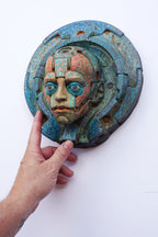 A hand holding a circular, relief-style sculpture of a stylized face with blue, green, and orange patina details against a plain white background.