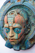 An ornate sculptural face mask or decorative headpiece with a circular halo-like frame, featuring turquoise, teal, coral, and beige painted details on a white background.