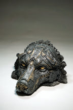A dark, highly detailed sculptural mask of a wolf with intricate metallic-looking gold patterns over a charcoal gray base, photographed on a neutral gray studio background.