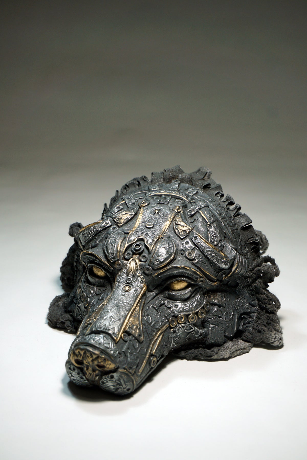 A dark, highly detailed sculptural mask of a wolf with intricate metallic-looking gold patterns over a charcoal gray base, photographed on a neutral gray studio background.