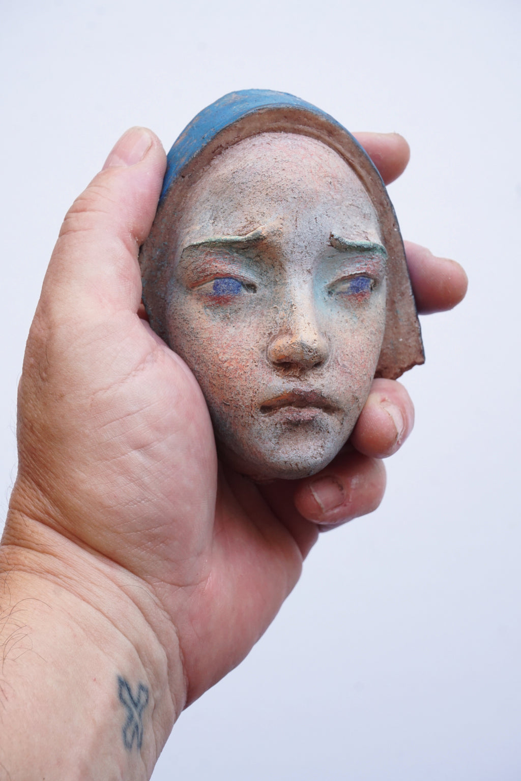 A hand holding a small sculpted ceramic or terracotta head with a blue head covering and pale blue eyes against a plain light background.