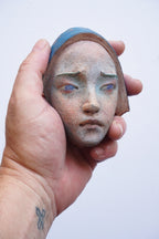 A hand holding a small sculpted ceramic or terracotta head with a blue head covering and pale blue eyes against a plain light background.