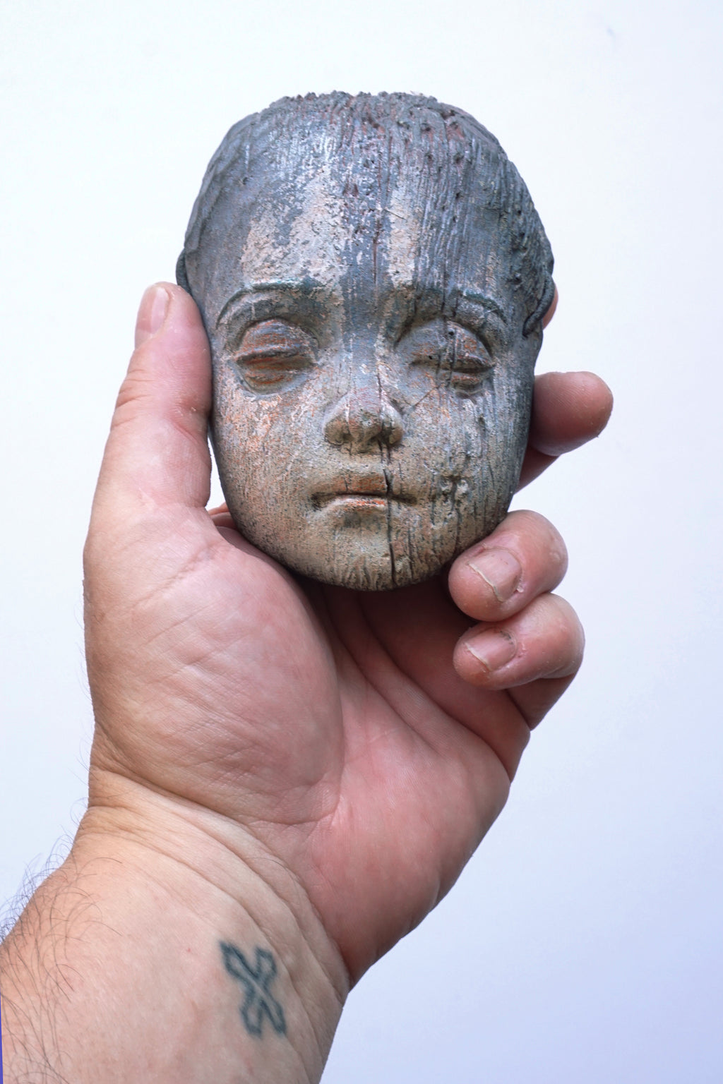 A hand holding a small carved wooden doll head with closed eyes, shown against a plain light background.