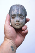 A hand holding a small carved wooden doll head with closed eyes, shown against a plain light background.