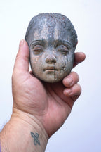 A hand holding a small carved wooden doll head with closed eyes, shown against a plain light background.
