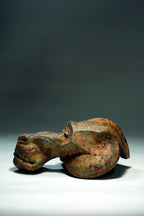 An ancient-looking mandril figurine with a weathered, rusted metallic surface showing brown, orange, and green patina, photographed on a white surface against a dark gray background.
