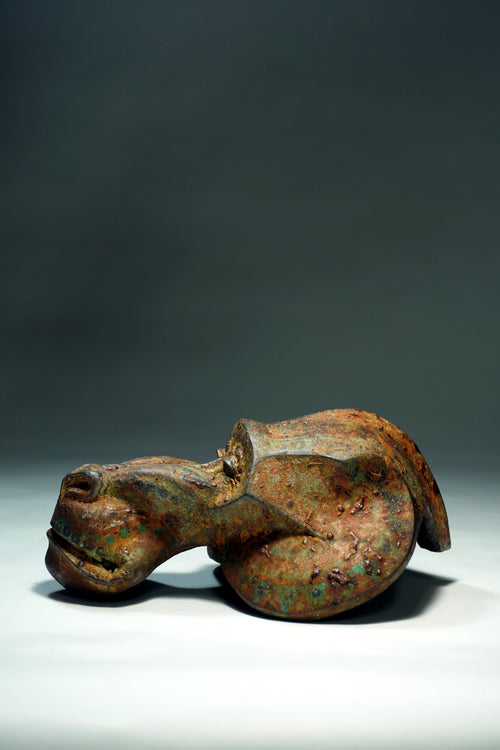 An ancient-looking mandril figurine with a weathered, rusted metallic surface showing brown, orange, and green patina, photographed on a white surface against a dark gray background.