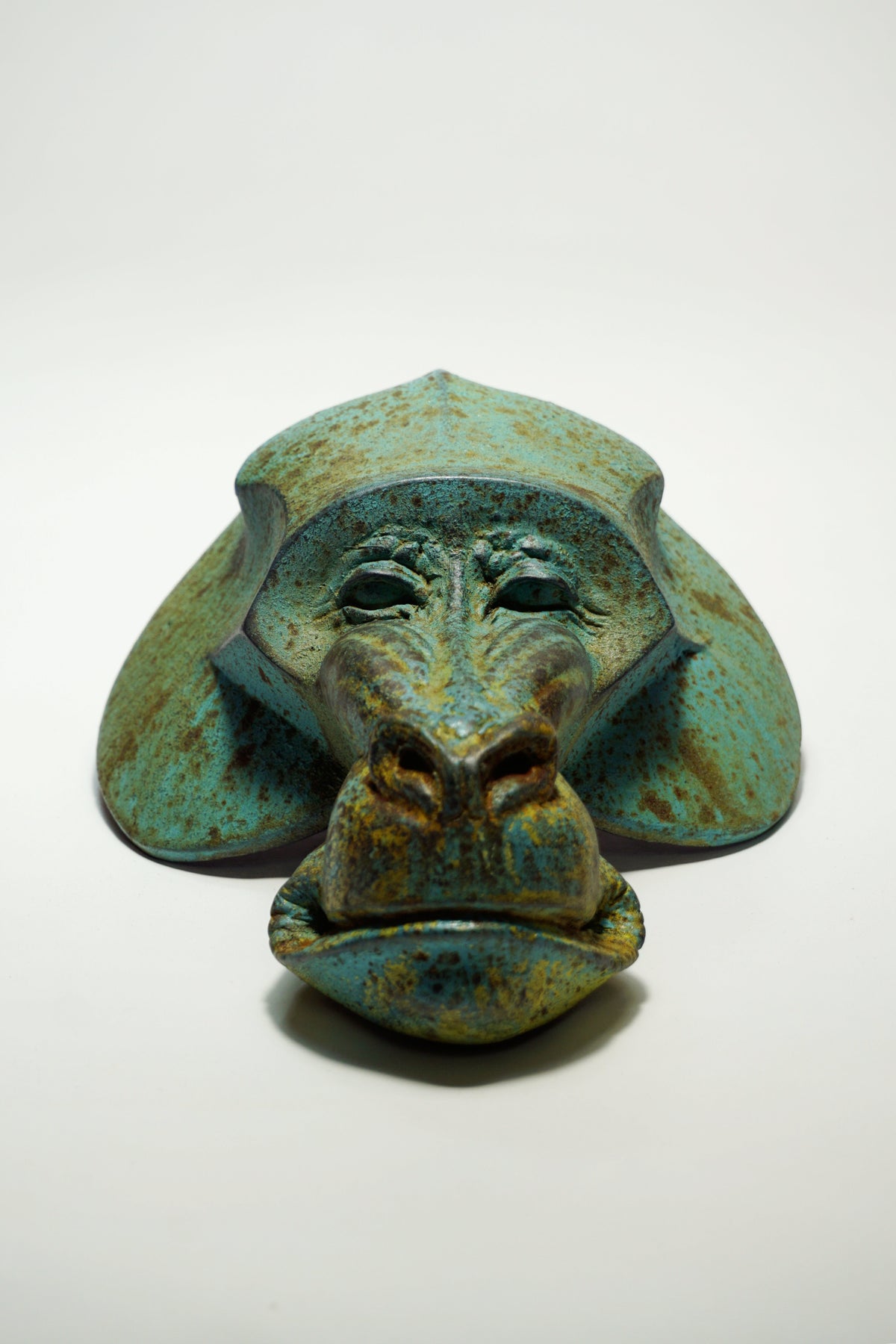 A sculptural monkey head mask or decorative object with a distressed turquoise, green, and brown patina finish, photographed on a plain white studio background.