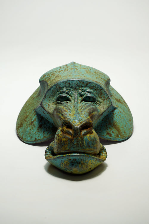 A sculptural monkey head mask or decorative object with a distressed turquoise, green, and brown patina finish, photographed on a plain white studio background.