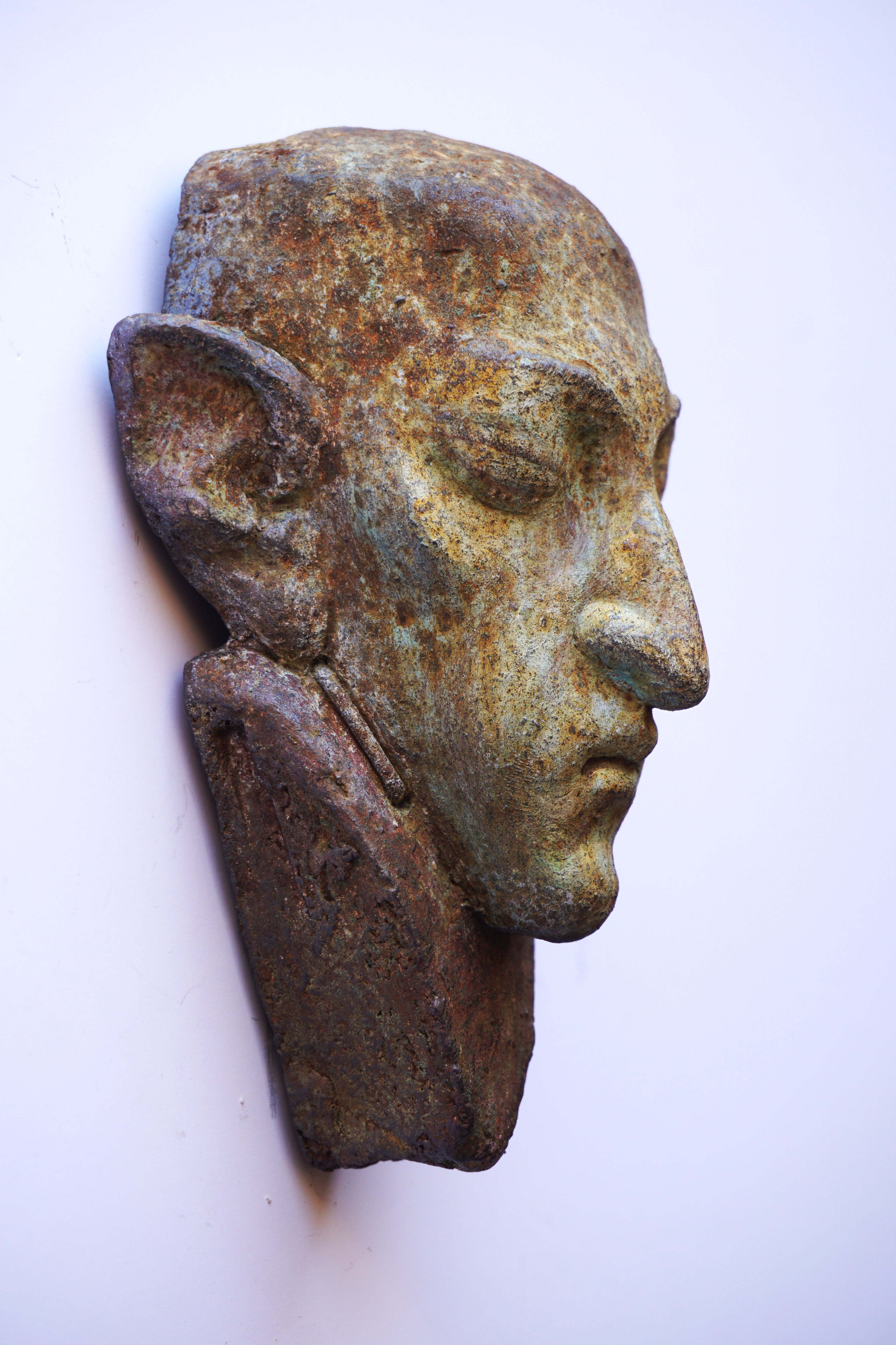 An antique-style metal head wall sculpture of a man's face in profile, featuring a textured patina with bronze, brown, and greenish hues, mounted against a plain off-white background.