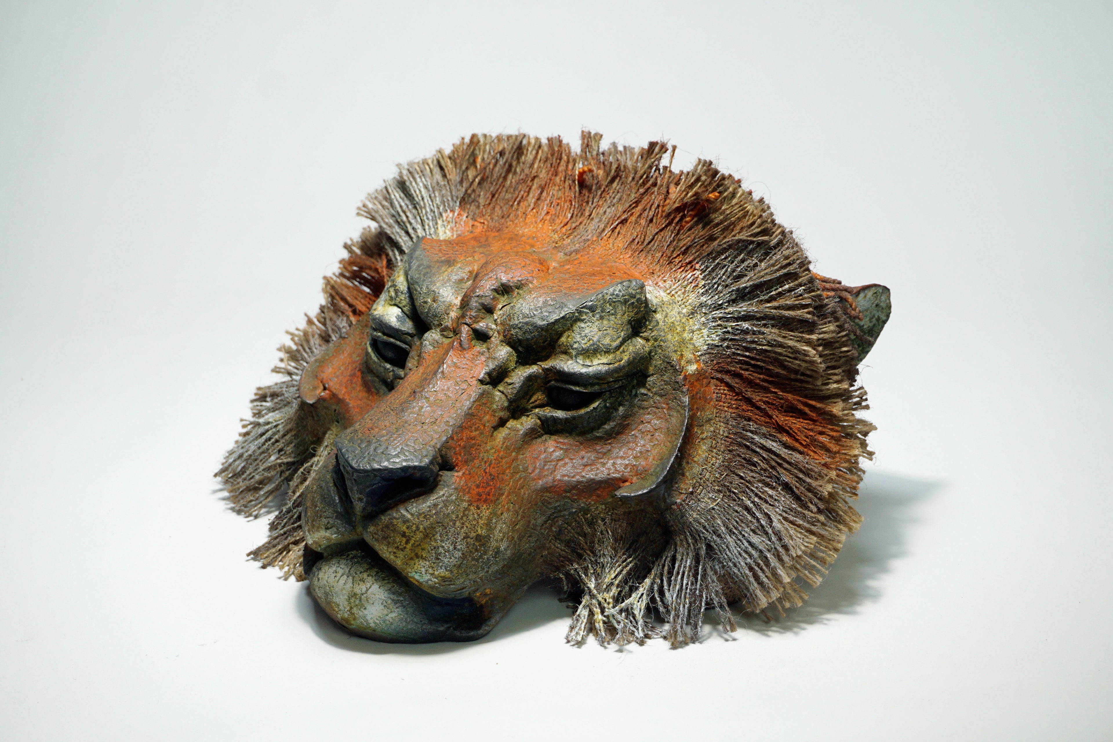 A sculptural lion head mask or decorative sculpture with textured fur details, shown on a plain light gray studio background.