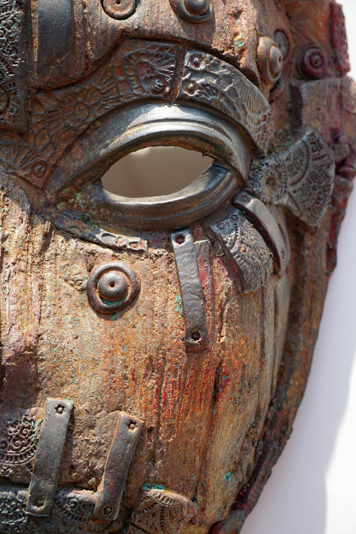 Close-up of a highly textured, rustic metal-like mask with intricate embossed patterns, weathered bronze and brown tones, and a central eye cutout.