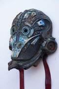 An intricate, hand-carved wooden mask with blue, white, and brown painted details, featuring a dark textured base and red hanging ribbons, shown against a plain white background.