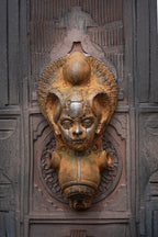An ornate bronze-toned metal door knocker featuring a stylized face with large ears and a halo-like top detail, mounted on a dark carved wooden door.