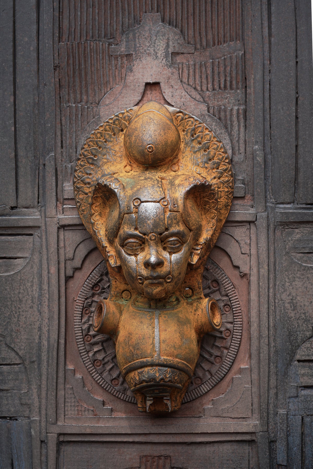 An ornate bronze-toned metal door knocker featuring a stylized face with large ears and a halo-like top detail, mounted on a dark carved wooden door.