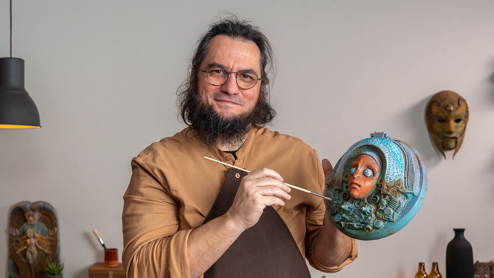 Man painting a mask in a studio setting with various masks on the wall.
